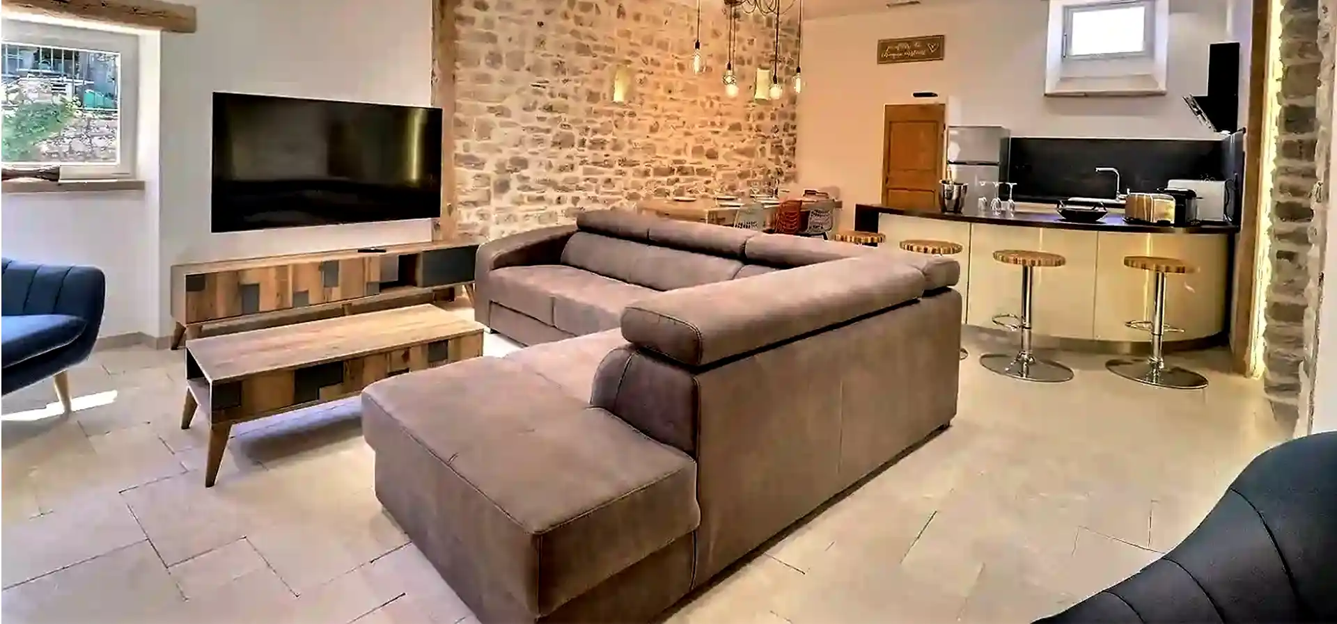 Salon avec grande TV 165cm et cuisine moderne tout équipée au Gîte Le Figuier à Gras avec table à manger et mur en pierre.